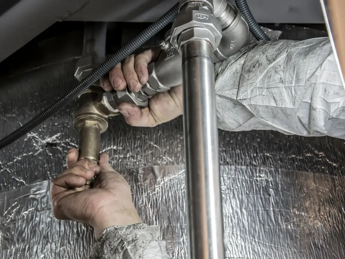 Plumber's hands tightening a brass fitting on overhead steel pipe beside foil-faced insulation in an Irvine, CA commercial building